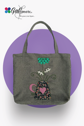 A gray handcrafted tote bag with a floral pattern and a green heart-shaped detail on a purple background, featuring a handle and a brand logo.