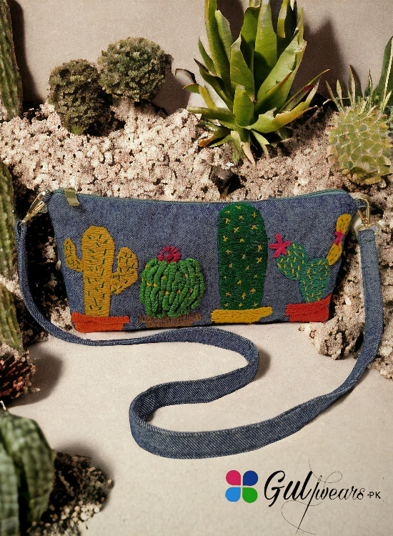 A grey clutch bag with 3D cactus embroidery, displayed against a backdrop of various cacti.