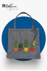 A gray tote bag with a handle, featuring a pattern of three cacti in different pots on the front, against a blue background.