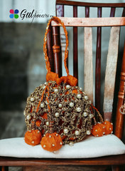 A bright orange potli bag with intricate copper gotta work, displayed on a wooden chair.