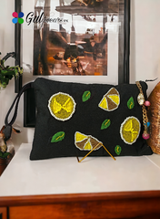 A black clutch bag embellished with pearl details and lemon and green embellishments, displayed on a table with a decorative background.