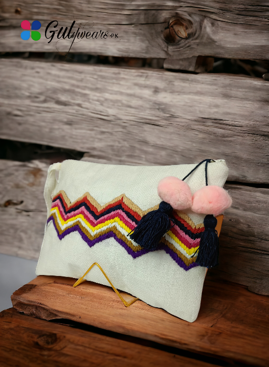 A white clutch bag with a handcrafted rainbow wave pattern and multicolored tassel hanging from the zipper.